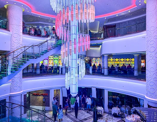 Vibrant and colorful atrium on the Norwegian Getaway cruise ship, featuring a striking multi-level chandelier, glass staircases, and illuminated casino slot machines in the background.