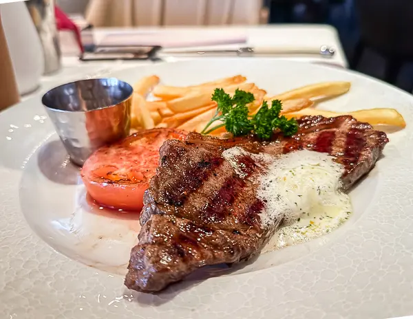 A grilled steak topped with herb butter, accompanied by crispy golden fries and a roasted tomato, served on a white plate at The Haven restaurant on Norwegian Getaway.