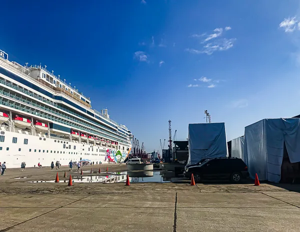 the Norwegian Pearl docked at the Santo Tomas de Castilla cruise port, which has an industrial setting. The area is open with concrete surfaces, a few scattered puddles, and some equipment covered with tarps. A few passengers are seen walking towards or from the ship.