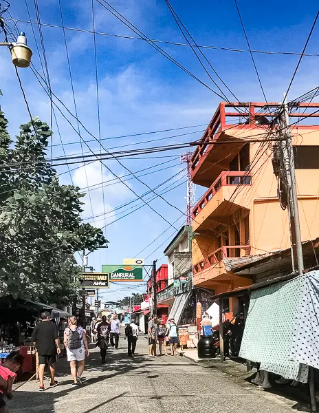 a street in Livingston, Guatemala, with several people walking along it. The street is lined with shops and has many overhead wires crisscrossing above. Part of the main street appears to be closed to vehicles.