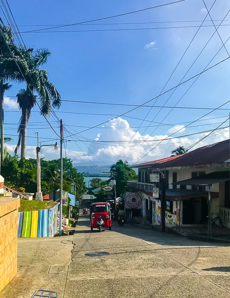 a tuk-tuk on a street in Livingston, Guatemala. The street is lined with buildings and overhead wires, with palm trees and a view of the mountains in the background.