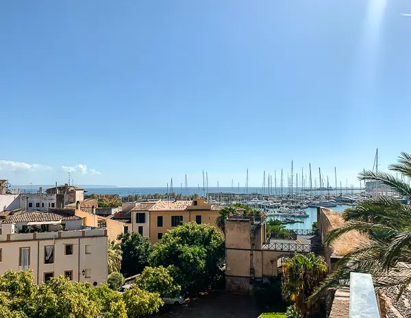 View over Palma de Mallorca's marina with sailboats docked in the harbor, surrounded by terracotta-roofed buildings, lush greenery, and a bright blue sky.