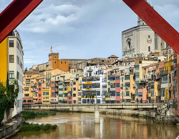 A view of Girona’s colorful houses along the Onyar River, framed by the red beams of the Eiffel Bridge, with the Girona Cathedral in the background.