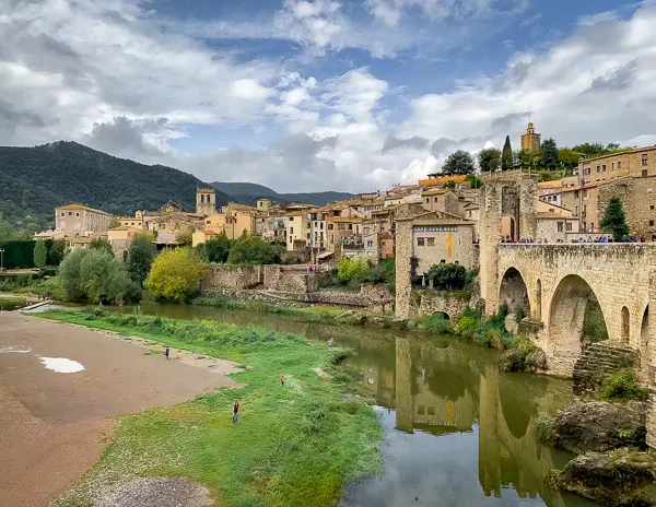 A panoramic view of Besalú, Spain, featuring its medieval bridge crossing the Fluvià River, with stone buildings and rolling hills in the background.