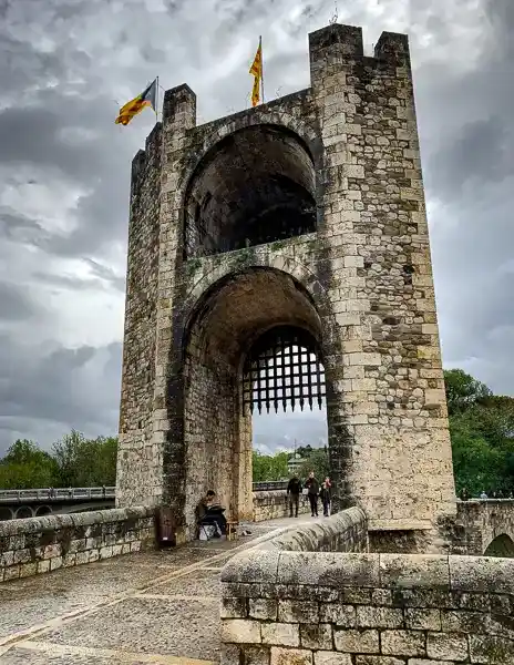 The entrance tower and medieval gate of Besalú, Spain, with stone arches, a raised portcullis, and Catalan flags waving above.