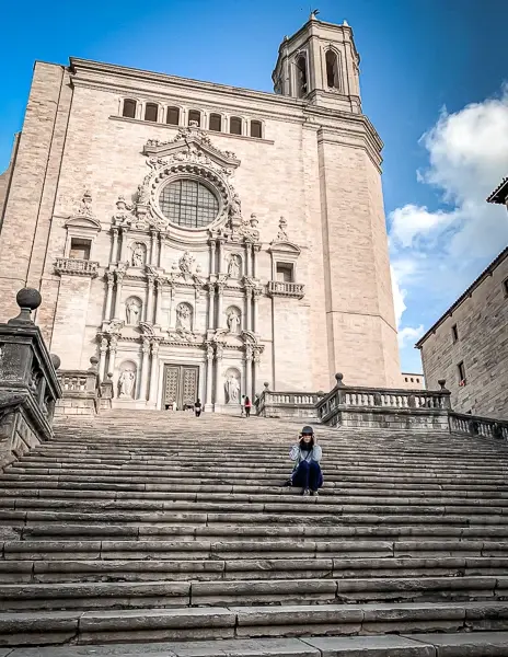 A person (kathy) sitting on the grand stone staircase leading up to the Girona Cathedral in Spain, with its intricate Baroque facade standing against a bright blue sky.