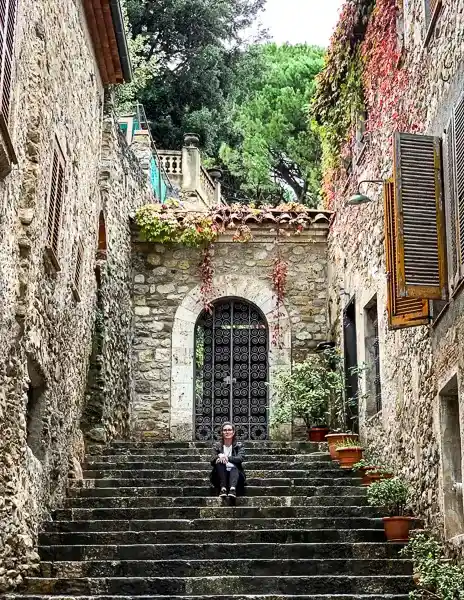 A person (kathy( sitting on stone steps in a charming medieval alley in Besalú, Spain, surrounded by ivy-covered stone buildings and rustic details.