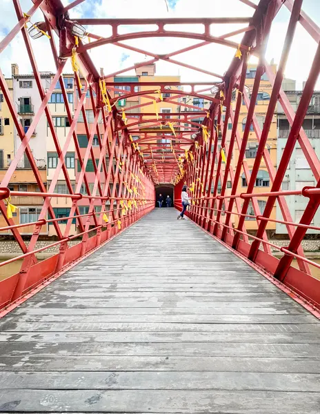 A view of the Eiffel Bridge (Pont de les Peixateries Velles) in Girona, Spain, with its red iron framework and yellow ribbons, leading toward the colorful buildings along the Onyar River.