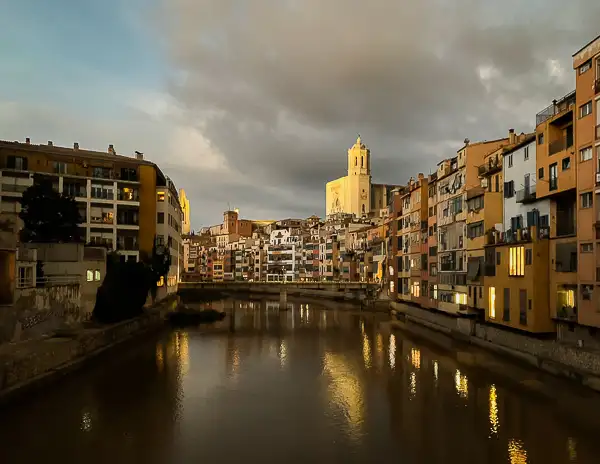 The Onyar River in Girona, Spain, at golden hour, reflecting the colorful riverside buildings with the Girona Cathedral standing in the background under a dramatic sky.