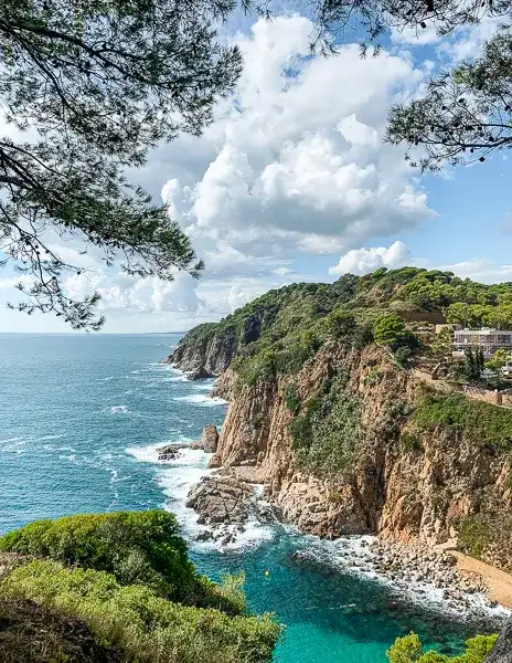 A breathtaking view of the rocky cliffs and turquoise waters of La Mar Menuda in Tossa de Mar, Costa Brava, Spain, framed by lush green trees and a bright blue sky.