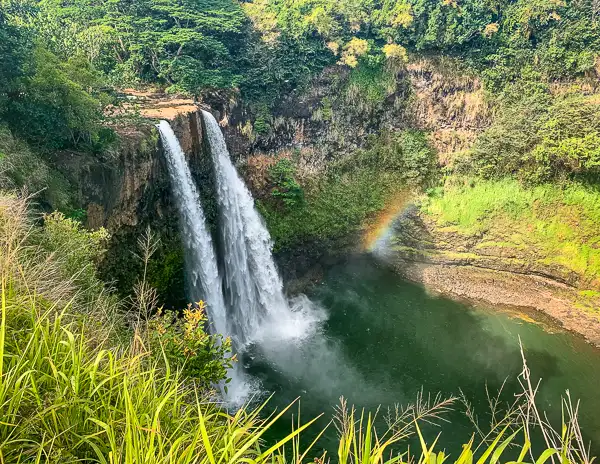 Wailua Falls in Kauai, a waterfall cascades into a serene pool, surrounded by lush greenery, with a visible rainbow formed in the mist near the base.