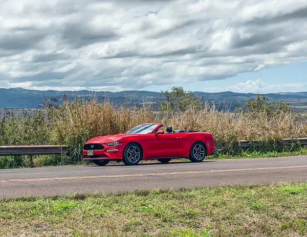 
A red convertible sports car parked beside a road in Kauai, with a backdrop of lush fields and distant mountains under a partly cloudy sky.