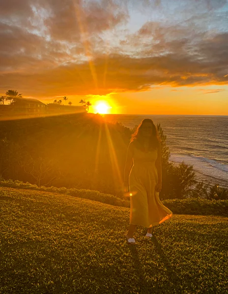 Kathy in a long dress stands on a grassy overlook at sunset, with the sun casting a brilliant glow over the ocean horizon behind her.