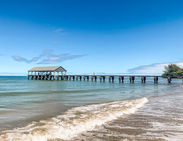 Hanalei pier with a sheltered area extends into calm, clear waters, set against a blue sky with few clouds.