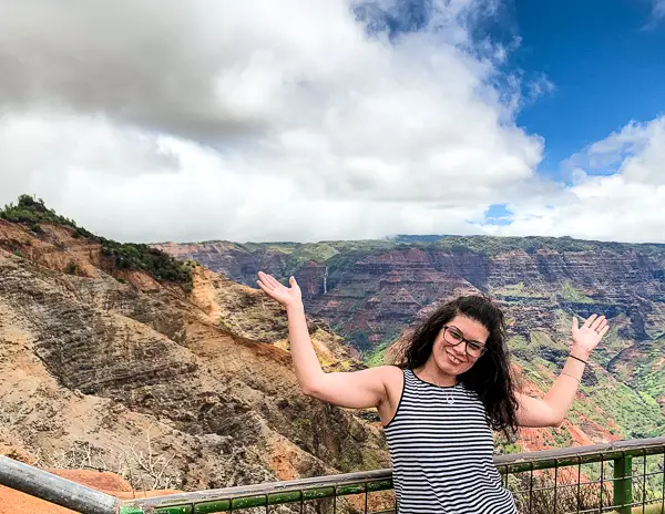 kathy happily poses with her arms raised, standing at a lookout with a panoramic view of Waimea Canyon’s layered red and green cliffs under a dynamic cloudy sky.