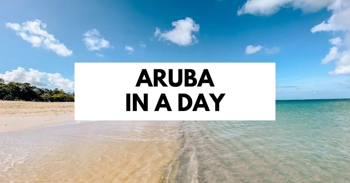 featured blog image features a serene beach scene with clear, shallow waters gently lapping onto a sandy beach, under a wide sky lightly dotted with clouds. The scene is framed with the bold caption "ARUBA IN A DAY" positioned centrally over a translucent white bar