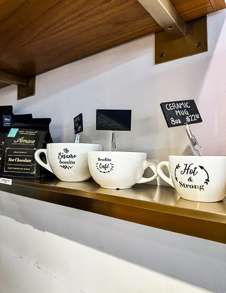 Merchandise display at Amara Café in Pasadena featuring ceramic mugs with playful designs and hot chocolate bags on a wooden shelf.