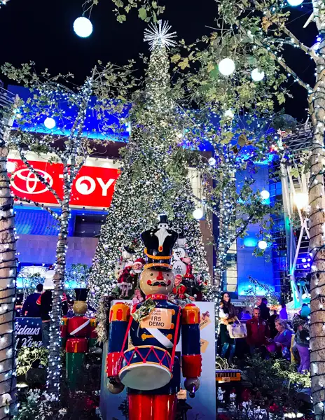 A giant Christmas tree illuminated with sparkling lights stands at LA Live, surrounded by festive nutcracker decorations and holiday displays.