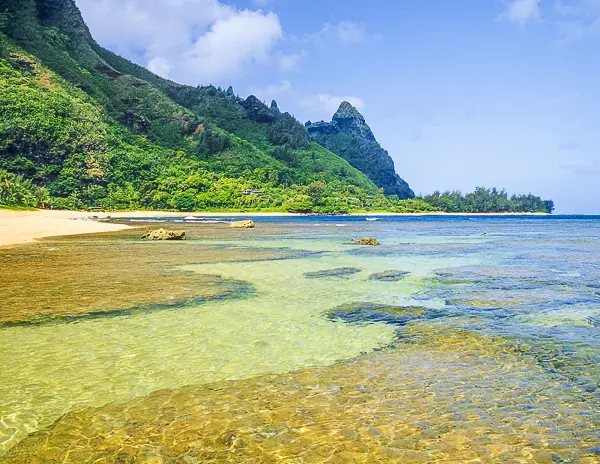 Tunnels Beach in Kauai with clear turquoise waters, a rocky shoreline, and lush green mountains in the background under a sunny blue sky.