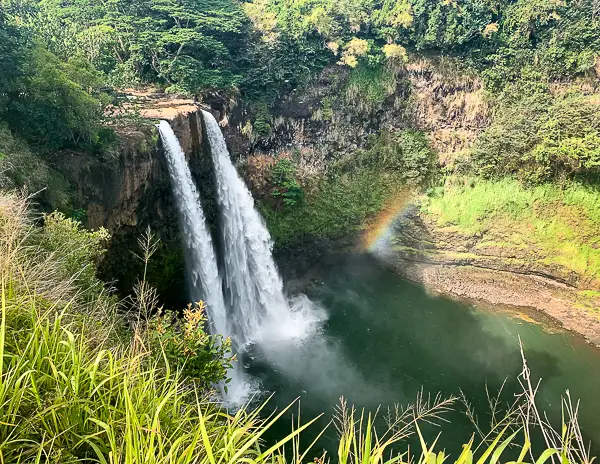 Wailua Falls in Kauai, Hawaii, cascading into a pool surrounded by lush greenery, with a vibrant rainbow visible in the mist.