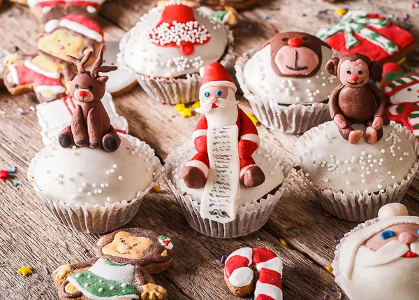 Festive cupcakes decorated with fondant Santa, reindeer, and other holiday characters, surrounded by decorated Christmas cookies on a rustic wooden table.