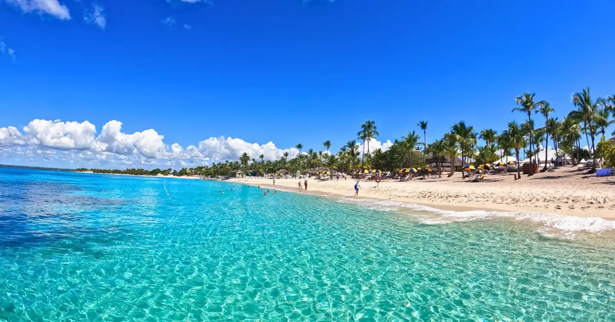 featured blog image of a stunning beach on Catalina Island, Dominican Republic, featuring clear turquoise water, white sand, and people relaxing under palm trees and umbrellas against a bright blue sky.