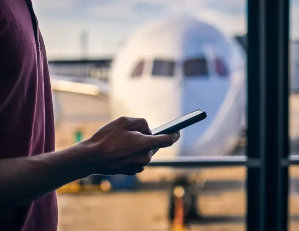 A close-up of a person holding a smartphone in an airport terminal, with a large airplane visible through the window in the background.