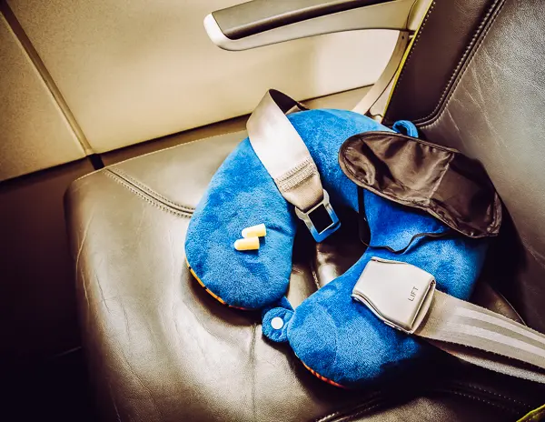 A blue travel neck pillow, an eye mask, and earplugs resting on a leather airplane seat, highlighting essential items for comfort during a flight.
