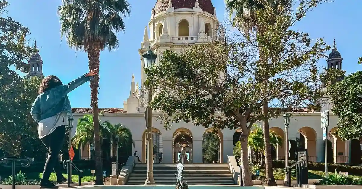 feaured image: A person standing in front of Pasadena City Hall, gesturing towards the iconic building with a fountain in the foreground and palm trees, representing exciting things to do in Pasadena.