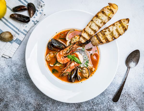 A bowl of cioppino garnished with herbs and served with grilled slices of bread, presented on a white plate with a striped napkin and a gray textured background.