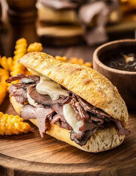 A French dip sandwich with melted cheese and roast beef on a crusty roll, accompanied by crinkle-cut fries and a small bowl of au jus for dipping, all served on a wooden board.