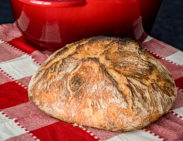A loaf of sourdough bread on a red and white checkered cloth, with a red pot in the background.