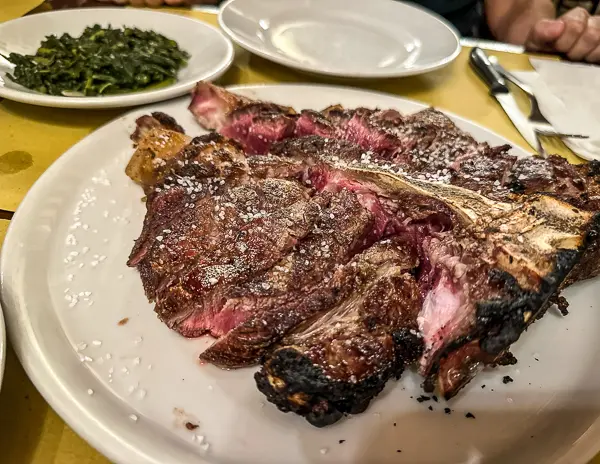 a beautifully cooked Bistecca alla Fiorentina, a Tuscan steak, served on a white plate, perfectly charred on the outside and pink inside, sprinkled with coarse salt. A side of cooked greens is visible on another plate in the background.