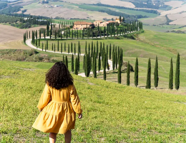 a person (kathy) in a yellow dress walking toward a picturesque Tuscan landscape with a winding road and cypress trees in Val