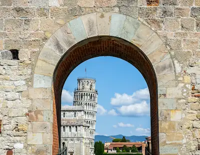 captures the Leaning Tower of Pisa framed by an ancient stone archway. The arch provides a unique perspective on the tower, which stands prominently against a backdrop of blue sky and distant hills