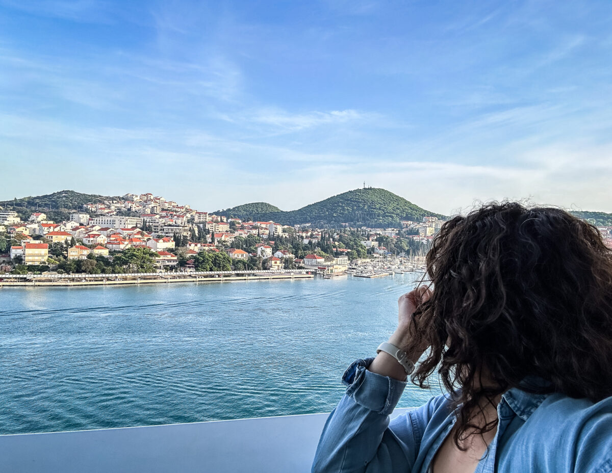 A person with curly hair (kathy), seen from behind, gazes out over the water from a cruise ship. The view features a coastal town in Croatia, with red-roofed buildings spread across rolling hills and a green, tree-covered mountain in the background.