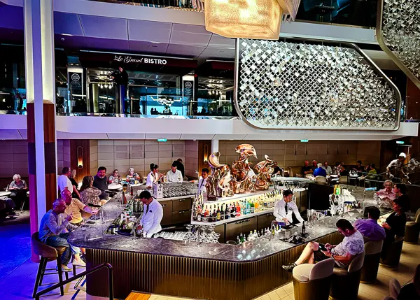 image is of the martini bar on celebrity beyond, a vibrant bar scene on what appears to be a cruise ship. At the center is a spacious, well-lit bar with bartenders actively mixing drinks. The bar features an impressive array of bottles and glasses, with patrons seated around, some interacting with the bartenders, others engaged in conversation or checking their phones. Above the bar, a stylish, modern lighting installation adds to the upscale ambiance. In the background, the upper level hosts a bistro, contributing to the multi-level, open-concept design of the venue.