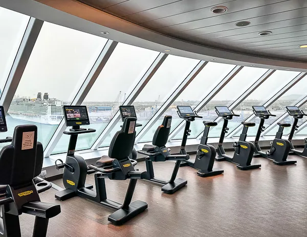 A row of stationary bikes in a cruise ship gym (Celebrity Beyond) with large windows providing a view of the port and another ship docked outside. The gym has a modern and clean design with each bike equipped with a digital screen.