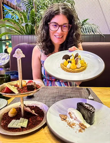 Kathy smiling while holding a plate of desserts, with more desserts displayed on the table in front.