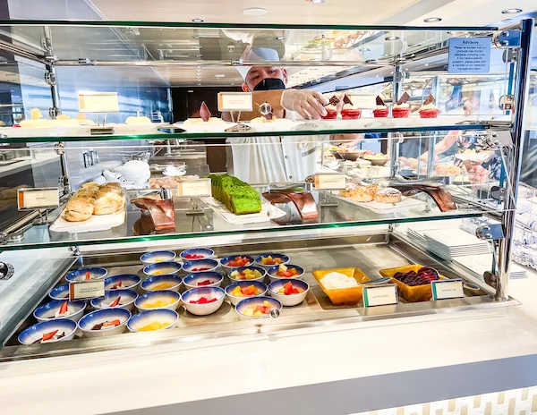 Chef arranging desserts on display at a cruise ship buffet, featuring a variety of pastries, cakes, and fruit bowls at Norwegian Joy's observation deck