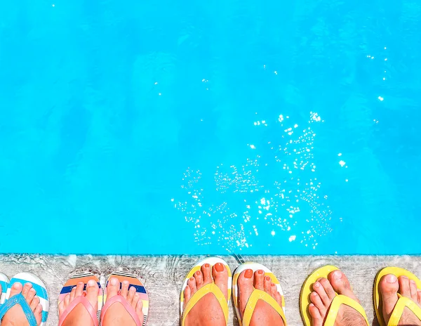 Close-up of feet in colorful flip-flops lined up by the edge of a pool.