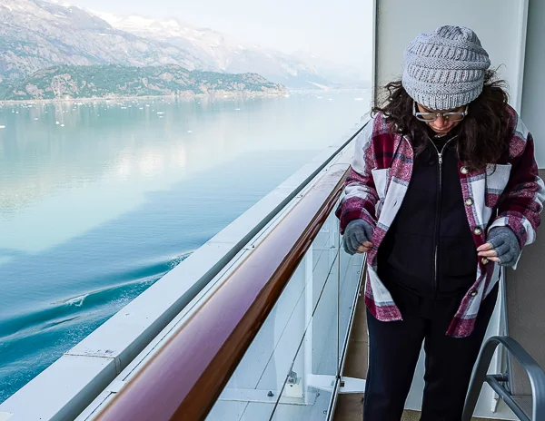 Kathy dressed in warm layers, including a beanie and plaid jacket, standing on a cruise ship balcony with scenic mountain views in Alaska.