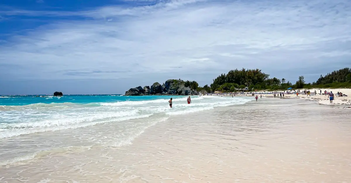 featured blog image without text of scenic view of a beach in Bermuda with turquoise waters and pink sand, where people are enjoying the shoreline under a partly cloudy sky. The beach extends towards rocky outcrops and lush greenery in the background