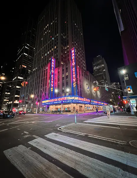 Corner view of Radio City Music Hall at night with neon lights glowing and city traffic passing by in Midtown Manhattan.