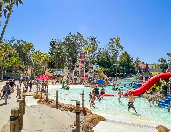 Vibrant scene at Splash Island in Raging Waters, featuring bustling water activities with children and families enjoying various slides and pools, surrounded by lush palm trees.