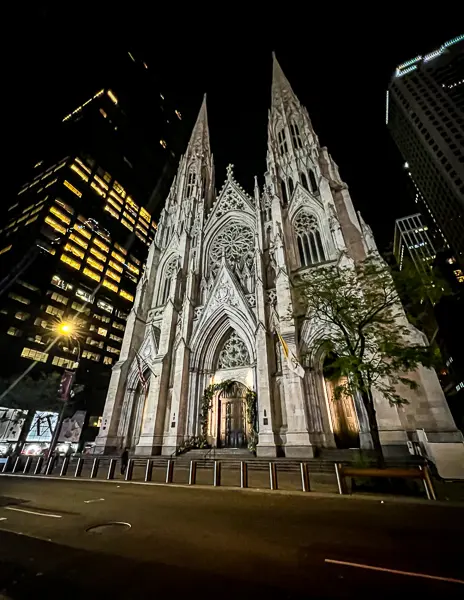 Nighttime photo of St. Patrick’s Cathedral in New York City, illuminated with gothic architecture standing tall among surrounding buildings.