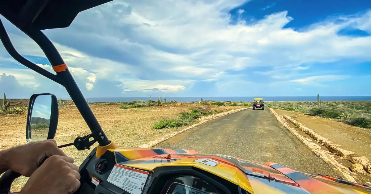 A view from inside an off-road vehicle, possibly a dune buggy or ATV, driving along a coastal road in Aurba with another similar vehicle in the distance. The landscape features arid terrain with scattered vegetation and cacti under a partly cloudy sky, indicating a rugged and adventurous setting after booking a shore excursion booked in Aruba.