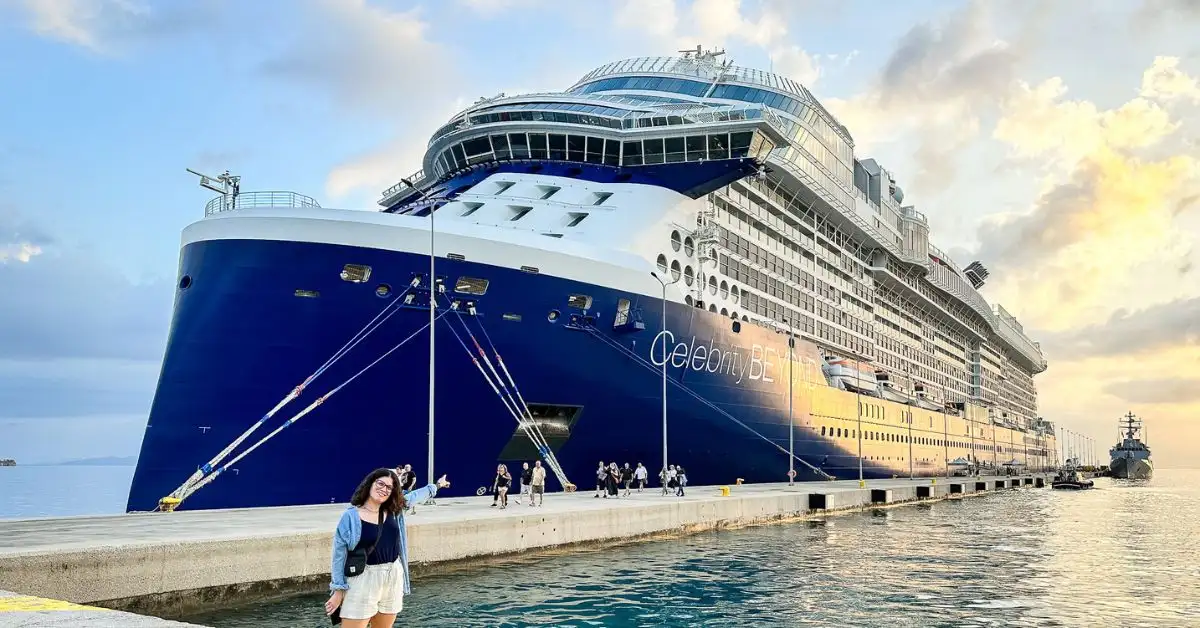 A woman standing on a dock in front of a large, blue cruise ship at sunset showing you how her cruise planning was successful/