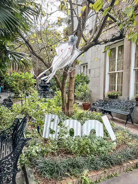 Halloween decoration in a New Orleans garden with a 'Boo!' sign and a ghost hanging from a tree.