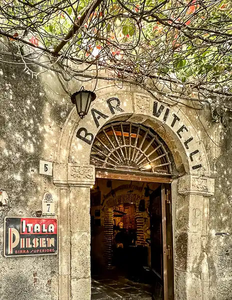 Charming entrance of Bar Vitelli, a picturesque location nestled under lush foliage. The archway is adorned with the bar's name and a vintage beer sign, leading into a cozy, inviting interior